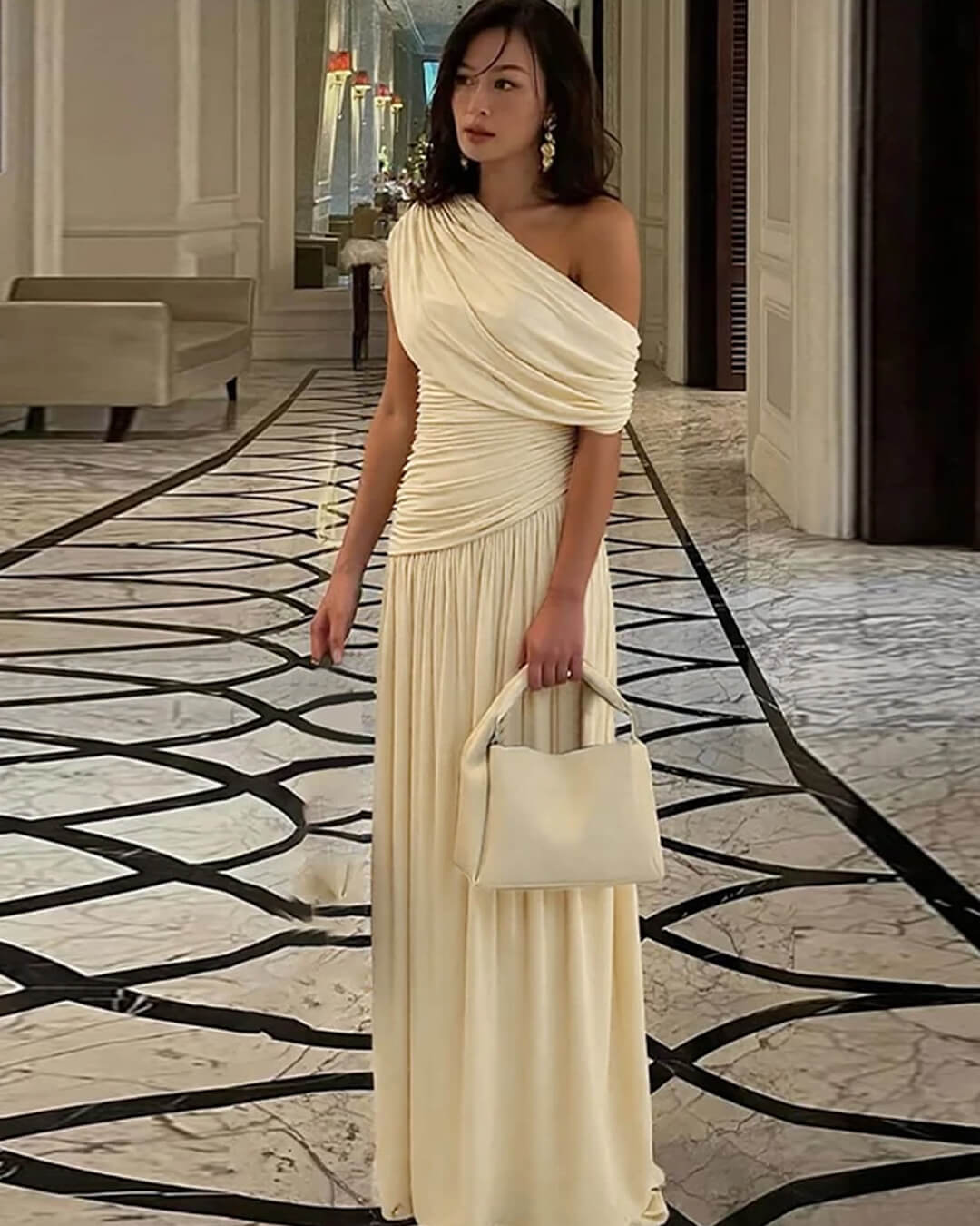 Robe longue drapée ivoire portée dans un hall en marbre avec sac à main