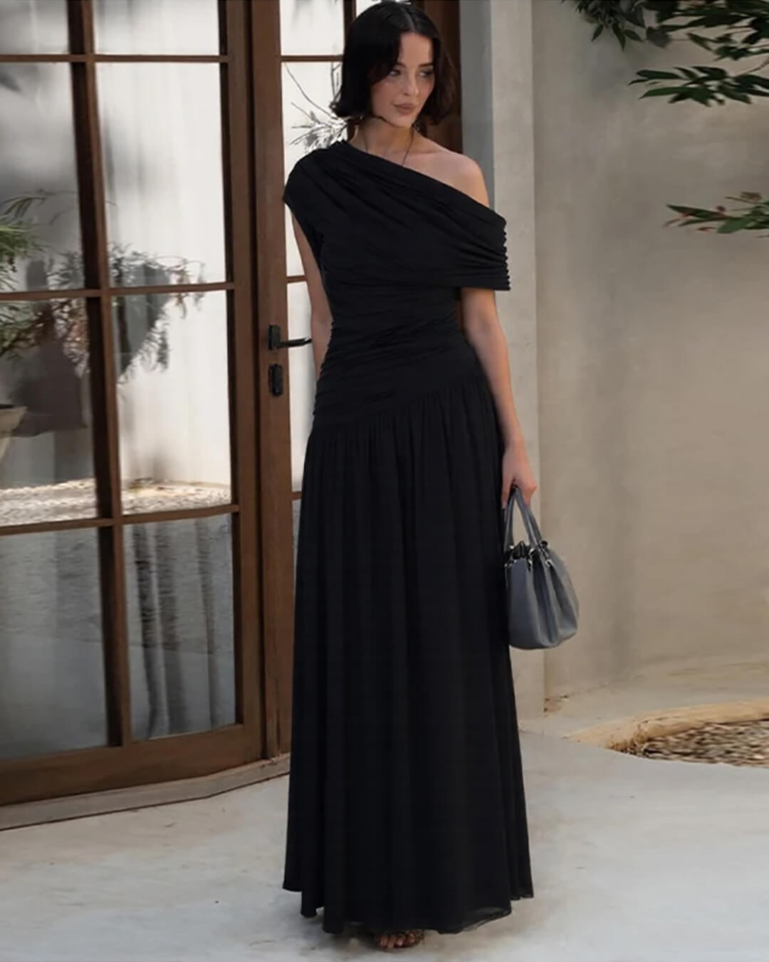 Robe longue drapée noire devant une porte vitrée, accessoirisée d'un sac